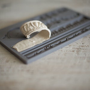May include: A gray silicone mold with a raised design of a decorative border. A white, flexible, silicone mold is placed on top of the gray mold.