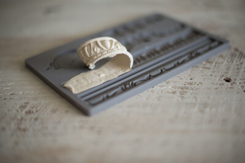May include: A gray silicone mold with a raised design of a decorative border. A white, flexible, silicone mold is placed on top of the gray mold.