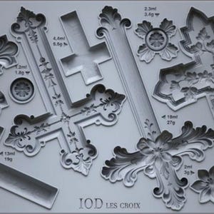 May include: A collection of gray, ornate cross-shaped molds and floral embellishments. The designs vary in size and detail, with measurements in milliliters and grams. The text "IOD LES CROIX" is visible.