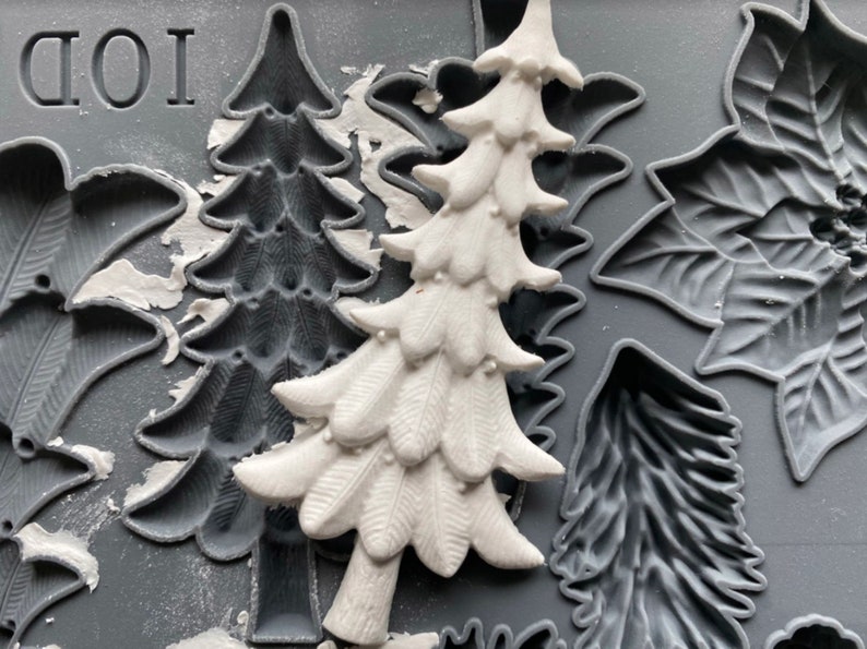 May include: A close-up of a silicone mold with various Christmas-themed designs. The mold features a tree, a poinsettia, and a leaf. The mold is gray and has a textured surface. The word "IOD" is printed on the mold.
