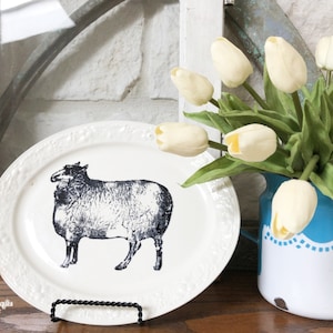 May include: White oval ceramic plate with a black and white illustration of a sheep. The plate is on a black metal stand. A blue and white enamel pitcher with a white lace trim holds a bouquet of white tulips.