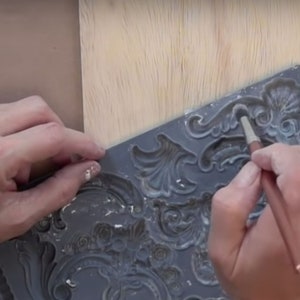 May include: A person using a brush to apply white paint to a gray silicone mold with a decorative design. The mold is used to create raised designs on surfaces.