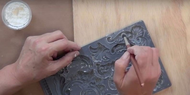 May include: A person using a brush to apply white paint to a gray silicone mold with a decorative design. The mold is used to create raised designs on surfaces.