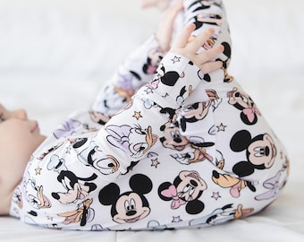 Op de afbeelding: Een witte baby-onesie met een herhaald patroon van Mickey Mouse, Minnie Mouse, Donald Duck en andere Disney-personages. De onesie met lange mouwen heeft een speels ontwerp met sterren en cartoonfiguren in zwart, roze en geel.