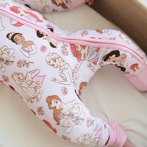 Può includere: Tutina rosa per neonato con cerniera e stampa di principesse Disney. La tutina ha polsini rosa e una cerniera rosa.