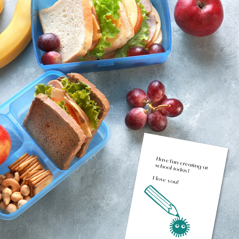 K&ouml;nnte beinhalten: Eine blaue Lunchbox mit einem Sandwich, Trauben und einem Apfel. Eine zweite Lunchbox mit einem Sandwich, N&uuml;ssen, Crackern und einem Apfel. Ein Zettel mit dem Text "Viel Spa&szlig; beim Kreieren in der Schule heute! Ich liebe dich!" und einer Zeichnung eines Bleistifts mit einem Keim an der Spitze.