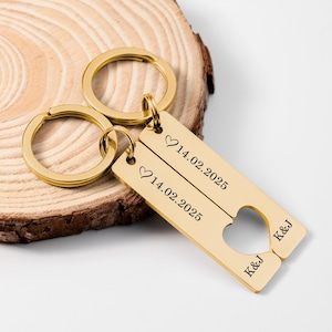 Peut inclure: Porte-clés en métal doré avec une découpe en forme de cœur. Chaque porte-clés comporte une étiquette rectangulaire avec la date "14.02.2025" et un symbole de cœur. Les initiales "K&J" sont également gravées sur les étiquettes. Les porte-clés sont sur une surface en bois.