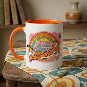 Peut inclure: Mug en céramique blanche avec une anse et un intérieur orange. Le mug présente un graphisme coloré d'un guépard sautant vers un arc-en-ciel, avec le texte "It's not all sunshine & rainbows but a good amount of it actually is."