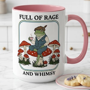 May include: White ceramic mug with a pink interior and handle. The mug features a whimsical illustration of a frog in a suit sitting on a mushroom, with the text "FULL OF RAGE AND WHIMSY".