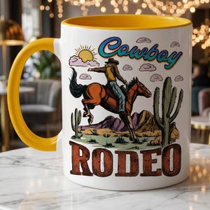 May include: White ceramic mug with a yellow handle and interior. The mug features a colorful illustration of a cowboy riding a horse in a desert landscape, with the words "Cowboy Rodeo" in a vintage font.