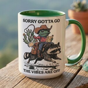 Peut inclure: Mug en céramique blanche avec intérieur et anse verts. L'illustration fantaisiste représente une grenouille cow-boy chevauchant un raton laveur, avec le texte "SORRY GOTTA GO" et "THE VIBES ARE OFF".