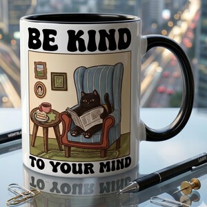 Peut inclure: Mug en céramique blanche avec intérieur et anse noirs. L'illustration représente un chat noir lisant un journal dans un fauteuil bleu, avec le texte "BE KIND TO YOUR MIND".