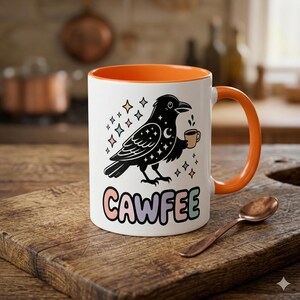 May include: White ceramic mug with an orange interior and handle. The mug features a black crow illustration holding a coffee cup, surrounded by stars. The word "CAWFEE" is printed in pastel colors below the crow.