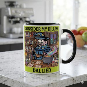Peut inclure: Mug en céramique blanche avec intérieur et anse noirs. L'illustration fantaisiste représente un opossum sorcier avec le texte "CONSIDER MY DILLIES DALLIED". Le design comprend un chaudron, un arc-en-ciel et des fioles de potion.