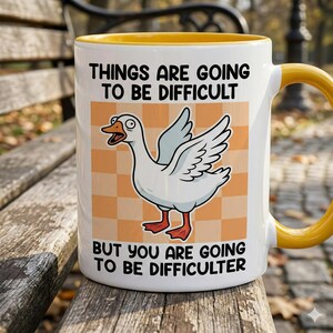 May include: White ceramic mug with a yellow interior and handle. The mug features a cartoon goose with text that reads, "THINGS ARE GOING TO BE DIFFICULT BUT YOU ARE GOING TO BE DIFFICULTER."