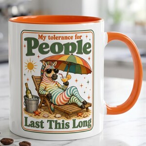 May include: White ceramic mug with an orange interior and handle. The mug features a cartoon zebra relaxing on a beach chair under a colourful umbrella, with the text "My tolerance for People Last This Long."