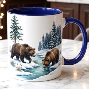 Peut inclure: Mug en céramique blanche avec intérieur et anse bleus. La tasse présente une scène hivernale avec deux ours bruns près d'une rivière enneigée et des arbres à feuilles persistantes. La tasse mesure environ 10 cm de haut.