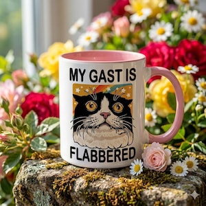 Peut inclure: Mug en céramique blanche avec intérieur et anse roses. Le mug présente un chat de dessin animé avec le texte "MY GAST IS FLABBERED" en noir. Le chat a des poils noirs et blancs et de grands yeux jaunes. Le mug est entouré de fleurs.