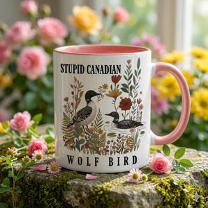 May include: White ceramic mug with a pink interior and handle. The mug features a design with two loons, floral elements, and the text "STUPID CANADIAN WOLF BIRD" in black lettering.