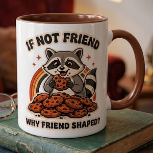May include: White ceramic mug with a brown interior and handle. The mug features a cartoon raccoon eating a cookie, with the text "IF NOT FRIEND WHY FRIEND SHAPED?" above and below the raccoon. A rainbow and cookies are also depicted.