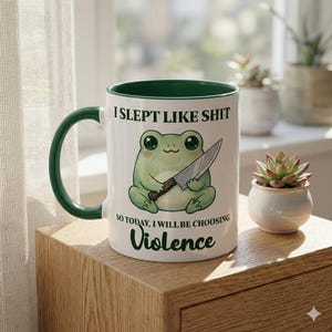 May include: White ceramic mug with a green handle and interior. The mug features a cartoon frog holding a knife, with the text "I SLEPT LIKE SHIT SO TODAY, I WILL BE CHOOSING Violence."