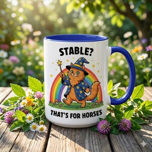 Peut inclure: Mug en céramique blanche avec intérieur et anse bleus. Le mug présente un chat de dessin animé habillé en magicien, tenant une baguette devant un arc-en-ciel. Le texte sur le mug se lit "STABLE? THAT'S FOR HORSES."