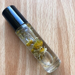 Op de afbeelding: Een transparante glazen rollerfles met een zwarte dop gevuld met een lichtgele vloeistof en gedroogde kamillebloemen.