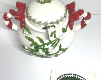 May include: A white ceramic sugar bowl with a lid, decorated with green leaves, white berries, and red bows. The handles are candy cane shaped. A green and white butterfly adorns the lid. The bowl is a festive holiday item.