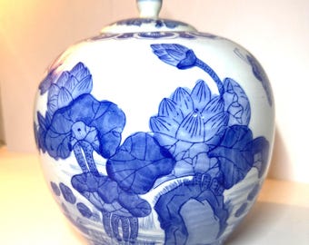 May include: A white ceramic jar with a lid, adorned with blue floral patterns. The jar has a round shape and a small, blue finial on the lid. The floral design features lotus flowers and leaves.