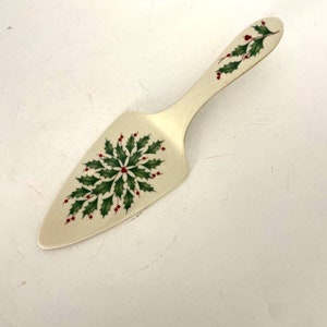 May include: A white ceramic cake server with a holly design. The server has a green holly leaf pattern with red berries.