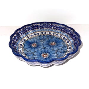 May include: A round ceramic dish with a scalloped edge, featuring a deep blue floral pattern with large blue flowers in the center and a decorative border of small blue and white geometric shapes and dots on a white background.