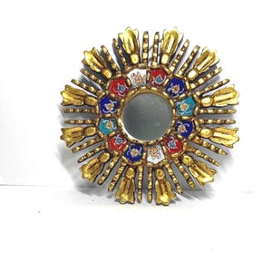 Puede incluir: Un espejo decorativo con forma de sol con marco dorado. El espejo tiene forma circular con una pequeña superficie reflectante. El marco presenta detalles de azulejos coloridos en rojo, azul y turquesa. El diámetro total del espejo es de aproximadamente 25 cm.