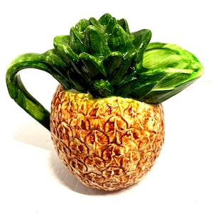 May include: A ceramic pineapple-shaped pitcher with a textured, brown body and a green leafy top. The handle is green and curved. The pitcher is designed to resemble a pineapple fruit.
