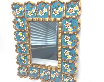 May include: Rectangular mirror with a gold frame. The frame has a repeating pattern of turquoise tiles with white and yellow floral designs. The mirror reflects a window. The frame is gold-coloured.