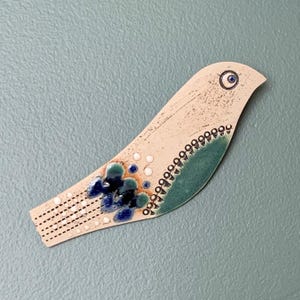 May include: A ceramic bird brooch in cream, with blue and teal details. It has a blue eye, a dotted border, and a tail with black dashes. A handcrafted, decorative accessory, suitable for adding a unique touch.
