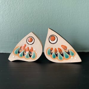 May include: Two triangular ceramic ornaments with a cream-coloured base. Each piece features a circular orange eye and a series of teardrop-shaped patterns in shades of orange, teal, and black, accented with small white dots along the bottom edge.