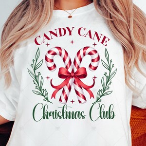 May include: White t-shirt with a Christmas-themed design. The graphic features two candy canes, a red bow, and the words "CANDY CANE" and "Christmas Club" in red and green. The shirt is a casual style.