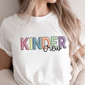 May include: White t-shirt with the text "Kinder Crew" in rainbow colors. The text is outlined in black.