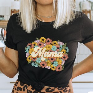 May include: A black t-shirt with a floral design and the word "Mama" in a retro font. The shirt is being worn by a person with blonde hair.