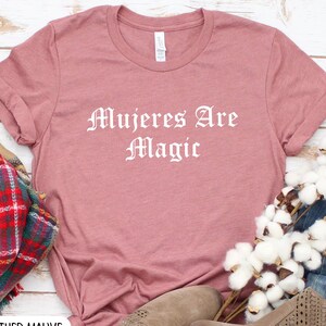 May include: Heather mauve t-shirt with the text "Mujeres Are Magic" in white lettering.