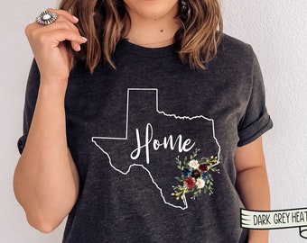 May include: Dark grey heather t-shirt featuring a white outline of the state of Texas with the word "Home" inside and a floral design in shades of red, blue, and white. The text "DARK GREY HEATHER" is visible at the bottom of the image.