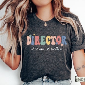 May include: A dark grey heather t-shirt with the text "DIRECTOR" in rainbow colors and "Mrs. White" in white script. The text is surrounded by small white stars.
