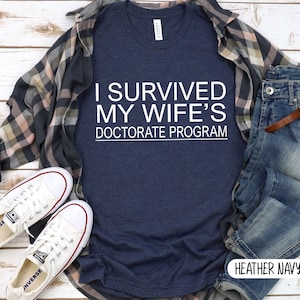 May include: A heather navy blue t-shirt with white text that reads "I survived my wife's doctorate program". The shirt is laid out on a wooden surface with a plaid shirt, jeans, and white sneakers.