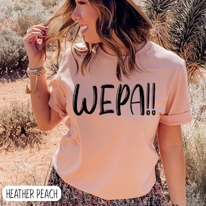 May include: A pink t-shirt with the text "WEPA!" printed in black on the front. The shirt is made of a soft, comfortable material and has a relaxed fit.