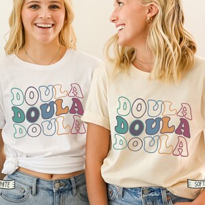 May include: Two women wearing white and cream t-shirts with the word "Doula" repeated in a retro font style. The shirts are available in white and soft cream.