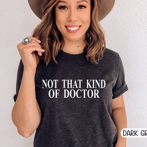 May include: Dark grey t-shirt with the text "NOT THAT KIND OF DOCTOR" in white. The shirt has rolled-up sleeves and is labeled "DARK GREY" in a white box. The model is wearing a hat and a ring.