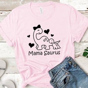 May include: Pink t-shirt with a black graphic of two dinosaurs, one larger than the other, with hearts and the text "Mama Saurus".