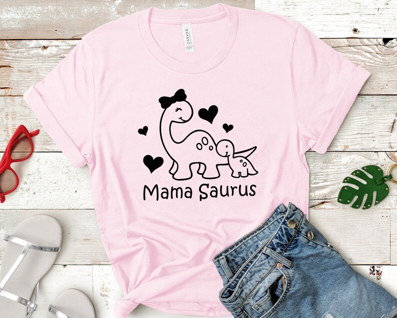 May include: Pink t-shirt with a black graphic of two dinosaurs, one larger than the other, with hearts and the text "Mama Saurus".