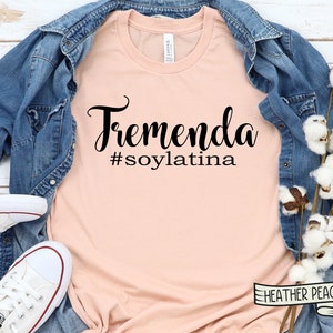 May include: A peach-colored t-shirt with the text "Tremenda #soylatina" in black lettering. The shirt is laid flat on a wooden surface with a denim jacket and cotton stems.