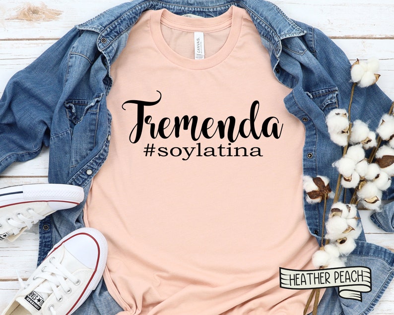 May include: A peach-colored t-shirt with the text "Tremenda #soylatina" in black lettering. The shirt is laid flat on a wooden surface with a denim jacket and cotton stems.